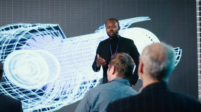 Black male speaker does manufacturing and engineering presentation with factory machinery. Businessman delivers pitch to mixed age audience at convention for industrial equipment. Camera B.