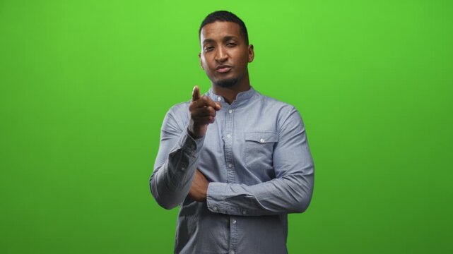 Young black man with arms crossed then points finger and gives thumbs up in a green screen studio; confidence approval.