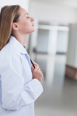 Woman standing in hospital corridor in lab coat holding stethoscope to chest, ring visible
