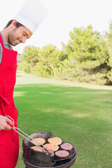 Indian man grilling burgers on charcoal grill using tongs wearing chef hat, red apron, copy space © wavebreak3