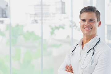 Male doctor posing arms-crossed smiling at clinic windows in lab coat, stethoscope, pen, copy space