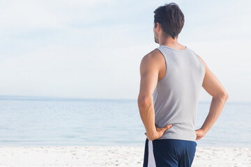 Man is standing on pale sandy beach wearing light gray tank top and navy swim shorts