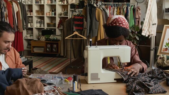 Medium pan shot of two multiethnic second hand outlet owners sitting together at table in store, African American woman sewing garment on machine, Caucasian man mending and upcycling preowned clothes