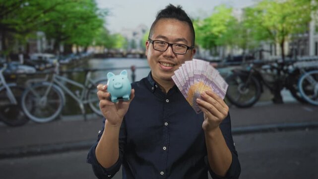 Young man smiling holding fan of rupiah banknotes and blue piggy bank on street in amsterdam; savings confidence.