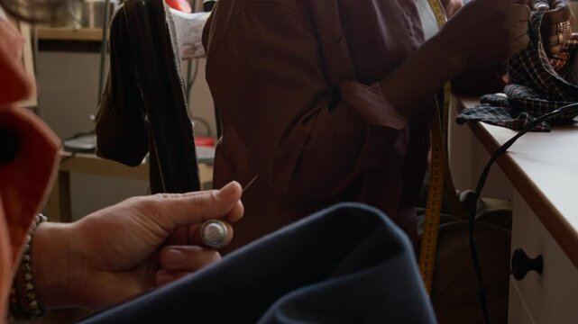Medium shot of two multiethnic young people upcycling preowned clothing in workshop at second hand store, man holding needle and thimble chatting to woman stitching on sewing machine