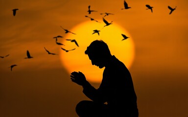  The silhouette of a man in prayer is set against a massive, glowing golden sun during a dramatic sunset. A flock of birds flies across the sky, adding a sense of freedom and life to the spiritual com