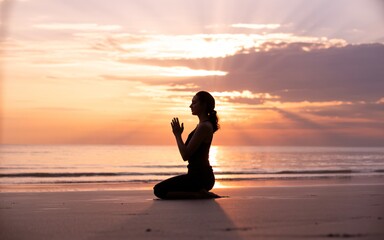  A woman sits in a peaceful meditative pose on a quiet beach as the sun sets over the horizon. The sky is filled with warm orange hues and soft light rays breaking through the clouds.