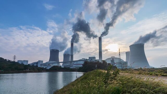 Time-lapse of a coal-fired power plant at early morning, cooling towers emitting steam against a soft sky, with calm water and green foxtail in the foreground.