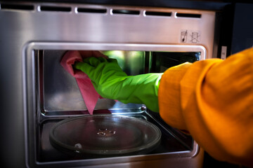 Hand in green glove scrubs corner of microwave oven with pink cloth. Kitchen appliance care, cleanup and domestic service concept