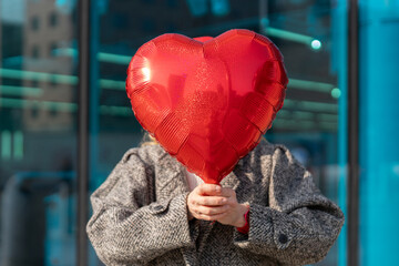 Holding a red heart balloon in front of a glass building on a sunny day with a person in a coat © Iryna