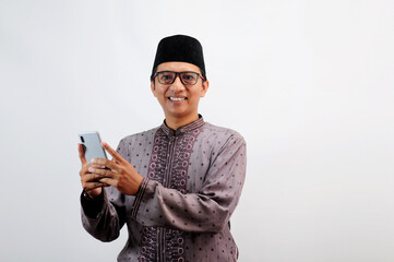 An Asian Muslim man smiles happily while sending a message using his mobile phone, isolated on white background