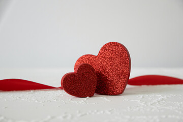 Two sparkling red glitter hearts stand together next to a vibrant red satin ribbon on a white surface.