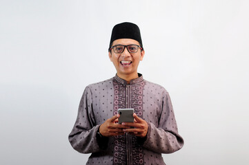 An Asian Muslim man smiles happily while sending a message using his mobile phone, isolated on white background