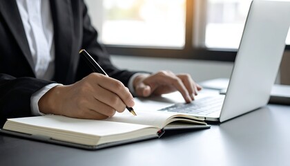 Person in suit writing in notebook, laptop open, near a window