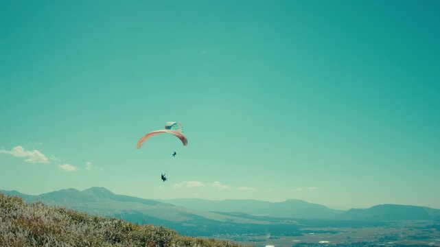 Two paragliders flying over green mountain landscape under blue sky, wide shot