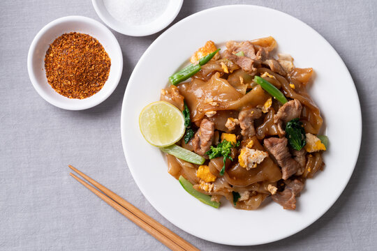 Top view of Thai food (Pad See Ew), Stir-fried rice noodles with soy sauce, pork, egg, and kale on a white plate, Served with fresh lime, chili flakes, and sugar