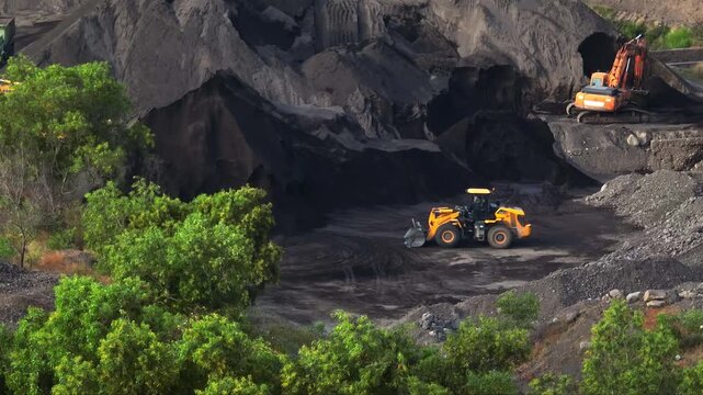 Heavy machinery including a bulldozer and an excavator operating in a large volcanic sand quarry extracting dark aggregate piles, surrounded by green vegetation
