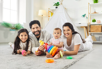 Portrait of smiling family of four with toys on rug at home looking at camera. Happy parents, older daughter and baby child playing with colorful stacking rings and creating joyful memories together © Studio Romantic