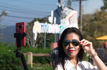 Beautiful Asian woman wearing sunglasses posing for a selfie using a red smartphone on a tripod in a sunny outdoor park, traveler vlogger lifestyle, professional photo editing in Photoshop