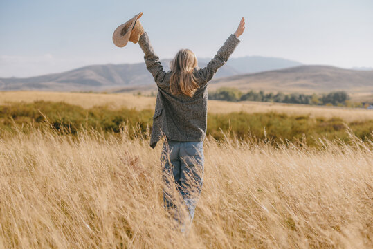 Blonde woman with raised arms holding hat in golden field at sunset. Back view of girl in Kazakhstan steppe with mountains. American west style, freedom and success concept. Warm autumn colors.