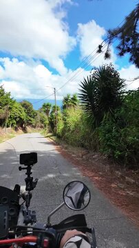 Point of view of a motorcyclist riding along a quiet mountain road surrounded by lush green vegetation under a blue sky with clouds. conveys freedom, adventure travel, road trip lifestyle, and outdoor