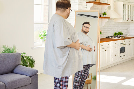 Obese overweight man examining mirror reflection. At home male person checks his belly, wearing glasses and a loose tee, contemplating health, body and acceptance. Obesity concept. 