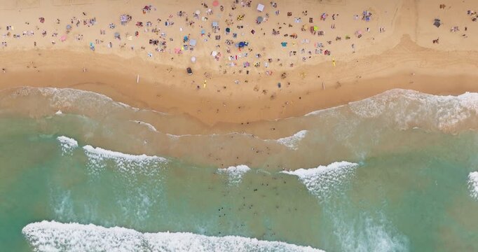 Bondi Beach shoreline full with people sunbathing on the sand and swimming in clear turquoise ocean water, featuring breaking waves. Summer holiday, travel, relax. Top down aerial drone shot zoom in