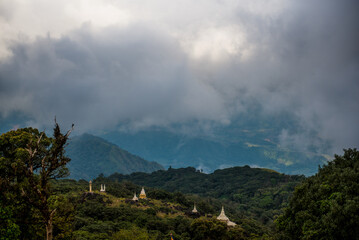 Scenic view of white Buddhist stupas on a lush green mountain ridge surrounded by thick morning mist and dramatic clouds in Southeast Asia.