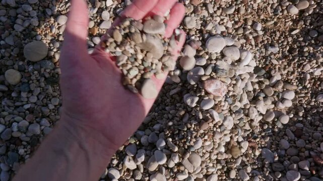 Point of view shot of human hand picking up small pebbles on pebble beach in sunlight, focusing on texture, relaxation, mindfulness and seaside experience.