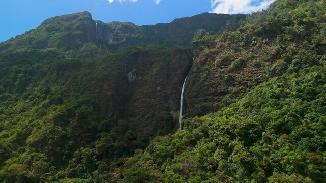 Cinematic drone flight moves toward the massive rocky face of El Chorro del Giron waterfall, highlighting the vertical cliffs, misty vegetation, and geological textures of the Azuay mountains