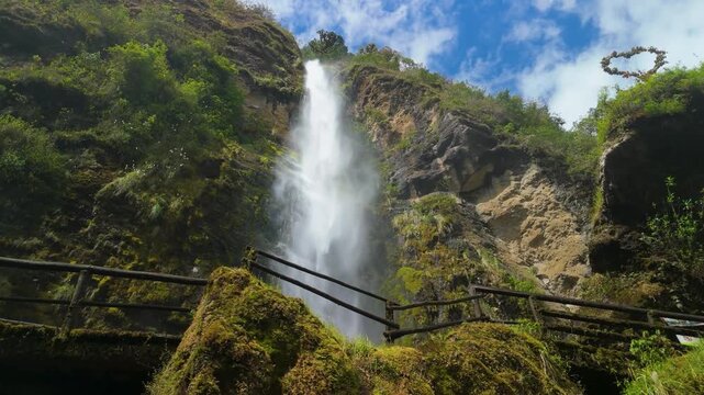 Spectacular parallax shot of powerful Chorro del Giron waterfall plunging from high rocky cliff surrounded by lush green vegetation and wooden railings under blue sky in Azuay province