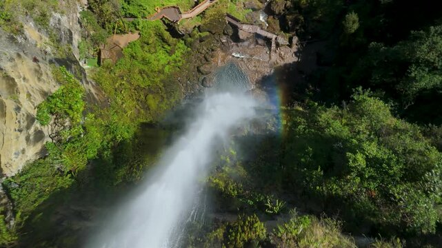 Dynamic aerial view plunging down powerful El Chorro del Giron waterfall stream toward rocky base and wooden bridge with vibrant rainbow appearing in mist against lush green cliffs of Azuay Ecuador