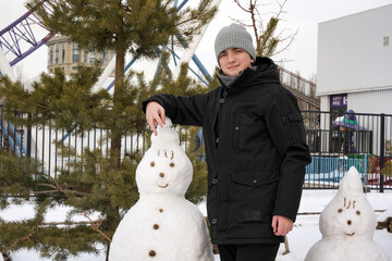 A young man (teenager) in a black jacket and grey hat stands next to a snowman in a snowy park....