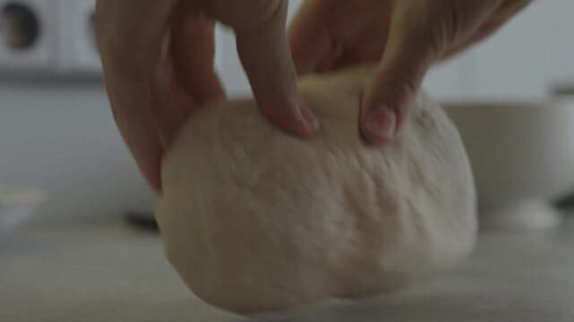 A person kneads dough on a countertop in a bright kitchen. The activity takes place in the morning. Hands work the dough to prepare for baking.