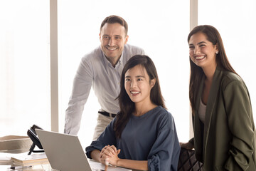 Fototapeta premium Diverse team of three happy coworkers meeting at laptop, standing and sitting at project manager workplace, looking away, smiling, enjoying collaboration, posing for candid group portrait