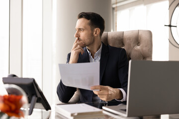 Naklejka premium Serious young Latin businessman thinking on marketing research results, looking away from workplace, holding paper sales reports, legal or financial documents, making decision, touching chin