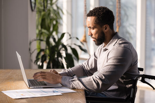 Focused African American business project manager man in wheelchair working at computer in office, typing on laptop, using online service, analyzing Internet marketing strategy. Side view shot