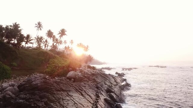The sun sets over the ocean, casting light on the rocky coast. Palm trees line the shore as waves crash against the rocks. The peaceful atmosphere invites relaxation and reflection.
