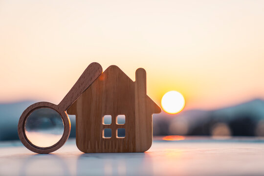 A wooden house model with a magnifying glass at sunrise, representing positive economic outlook, real estate opportunity, financial institution analysis, investment confidence, market recovery. 