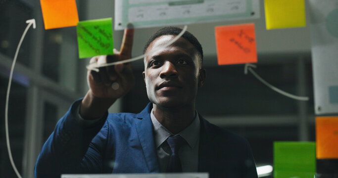 Glass wall, night or black man in office with review, strategy or planning for report. Problem solving, sticky note or employee with insight, idea board or brainstorming deadline on evening shift.