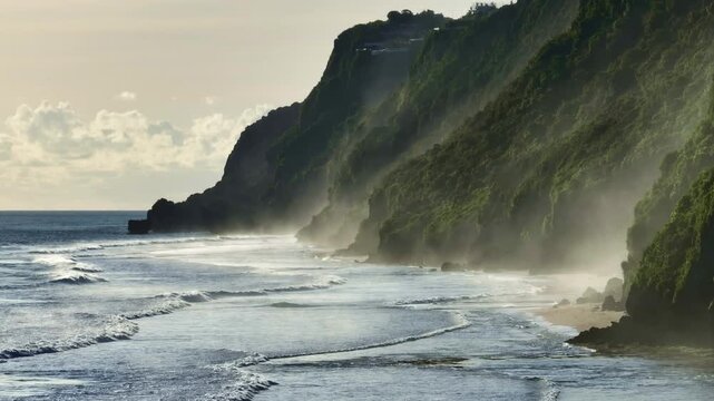 Bukit Peninsula coastline in Bali lush green cliffs rising from the Pacific Ocean, with waves breaking on shore and a light mist lingering along the tropical beachfront. Natural background static shot