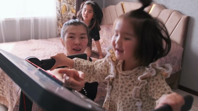 A toddler girl walks on treadmill at home under careful supervision of her mother, while her sisters watch and play nearby. Cozy family atmosphere and home fitness. Little girl walking on treadmill 