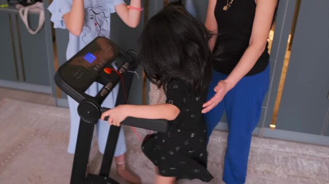 A young girl runs on treadmill at home, supported by her sisters and supervised by her mother for safety. Family fitness, healthy lifestyle, and childhood activity indoors. Little girl running on tr