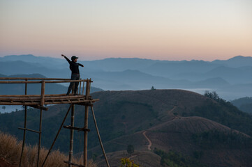 Naklejka premium A woman stands on a bamboo bridge in the early morning as the sun is rising, with mountains in the background.
