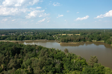 Fototapeta premium Expansive aerial landscape featuring a calm forest lake bordered by dense green woodland, with a rural village and open fields visible on the horizon under a bright blue sky with scattered clouds near