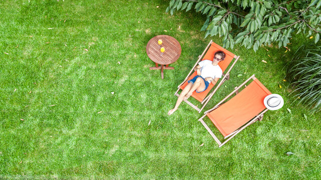 Young girl relaxes and reading book in summer garden in sunbed deckchair on grass, relaxing outdoors in green park on weekend, aerial drone view from above