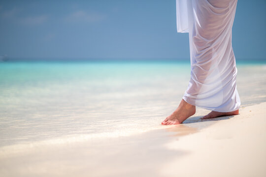 woman with bare legs in white sarong stands in calm blue sea, gently dipping her feet, evoking tranquility and peace, serenity, relaxation, escape., wellness, mindfulness, calm, serenity., None