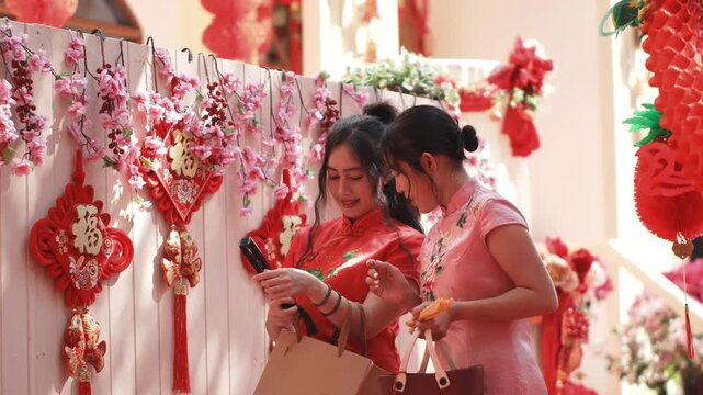 Two happy Asian women in Cheongsam dresses holding shopping bags and smiling during Chinese New Year celebration, medium eye-level shot