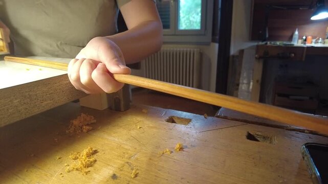 Close-up of a violin bow maker shaping a wooden bow stick on a workbench using a hand plane and wooden jig, showing traditional luthier craftsmanship and precision instrument making.