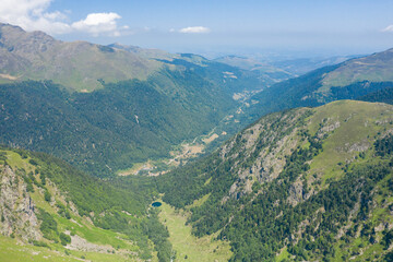 High-angle view of a lush green valley surrounded by steep forested mountains in the Pyrenees, with rolling hills fading into the horizon under a bright summer sky.
